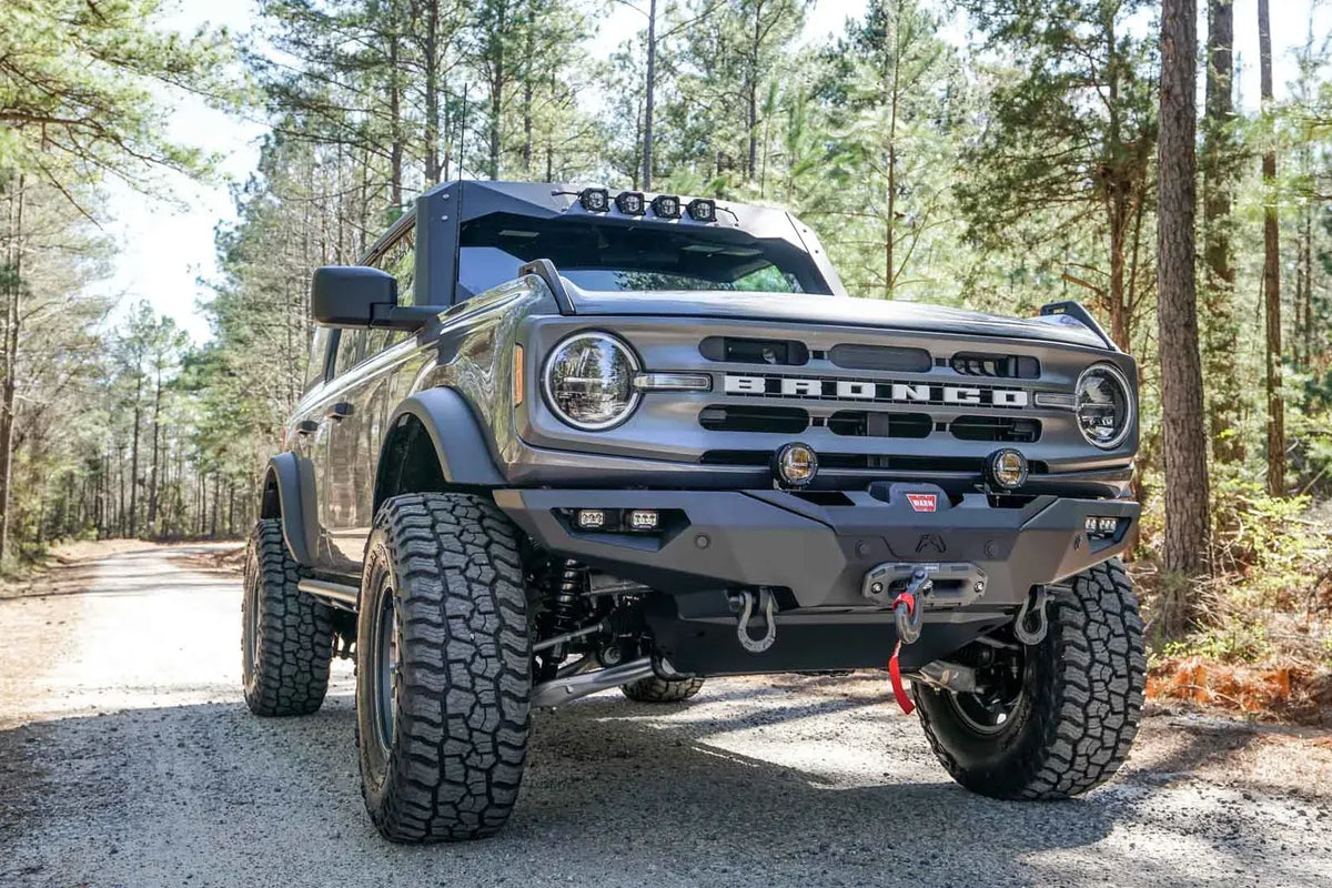 Fab Fours Lifestyle winch front bumper on a lifted gray Ford Bronco on a gravel road in the woods, passenger-side front three-quarter view, with Warn winch and D-ring shackles
