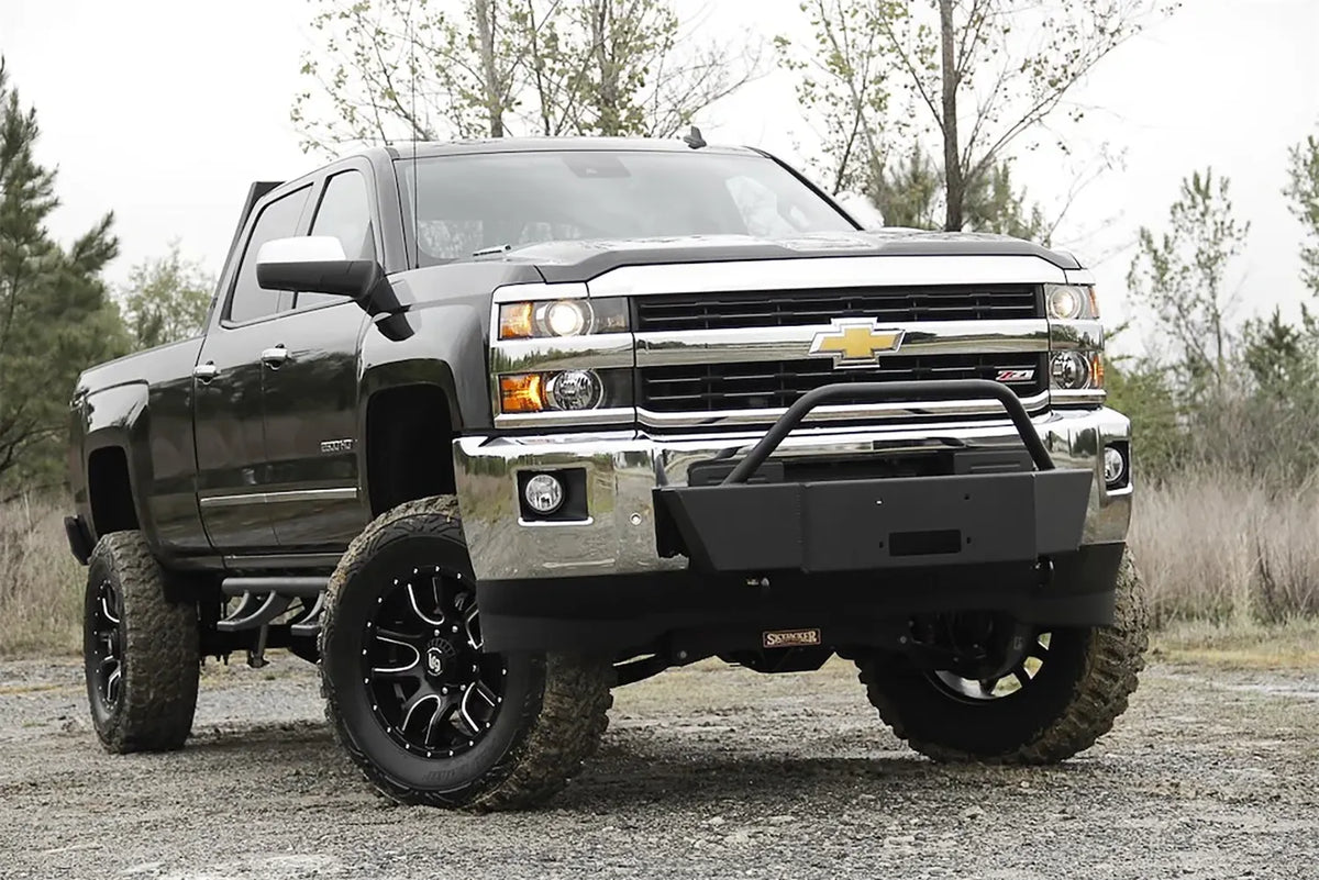 Fab Fours winch mount with pre-runner guard on a black Chevrolet Silverado 2500HD parked on gravel, front passenger-side three-quarter view, showing the pre-runner hoop, winch plate and factory bumper integration