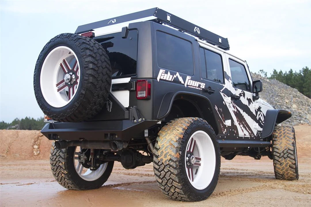 Fab Fours heavy-duty rear bumper with tire carrier on a lifted black and white Jeep Wrangler JK Unlimited with Fab Fours graphics and roof rack, passenger-side rear three-quarter view on a dirt lot