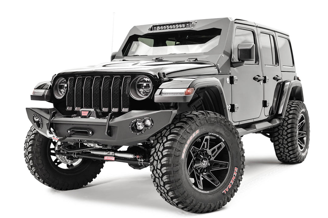 Fab Fours Lifestyle winch bumper on a gray Jeep Wrangler JL, driver-side front three-quarter view, showing the bumper with winch, red recovery hook strap and light pods