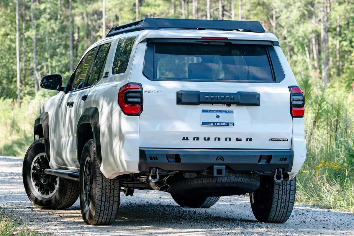Fab Fours Premium rear bumper on a white Toyota 4Runner on a gravel road in the woods, driver-side rear three-quarter view, with D-ring shackles