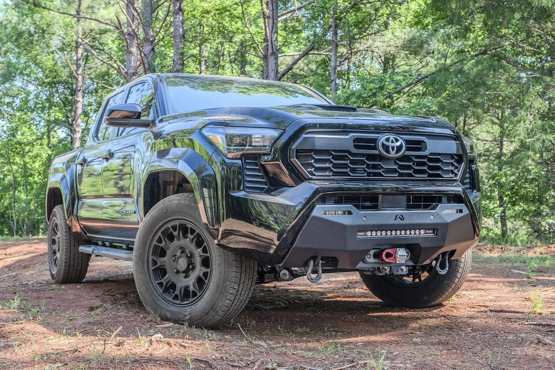 Fab Fours Hidden Winch Mount on a black Toyota Tacoma, front three-quarter view outdoors, showing the winch fairlead, LED light bar, and D-ring shackles