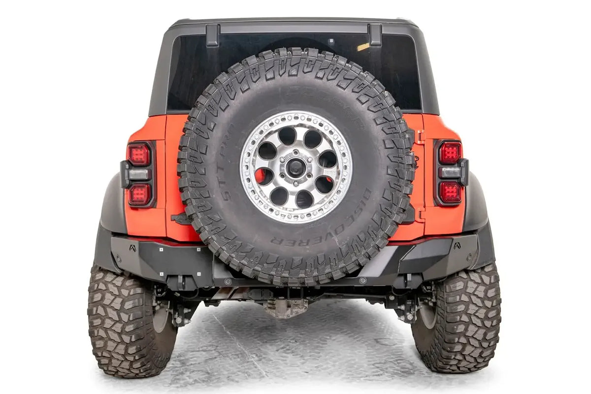 Matte black Fab Fours Premium rear bumper on an orange Ford Bronco Raptor, straight rear view, with parking sensors, license plate area, and tailgate-mounted spare tire