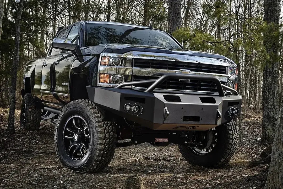 Fab Fours Premium heavy-duty winch front bumper with pre-runner guard on a lifted black Chevrolet Silverado 2500HD, passenger side, parked in the woods