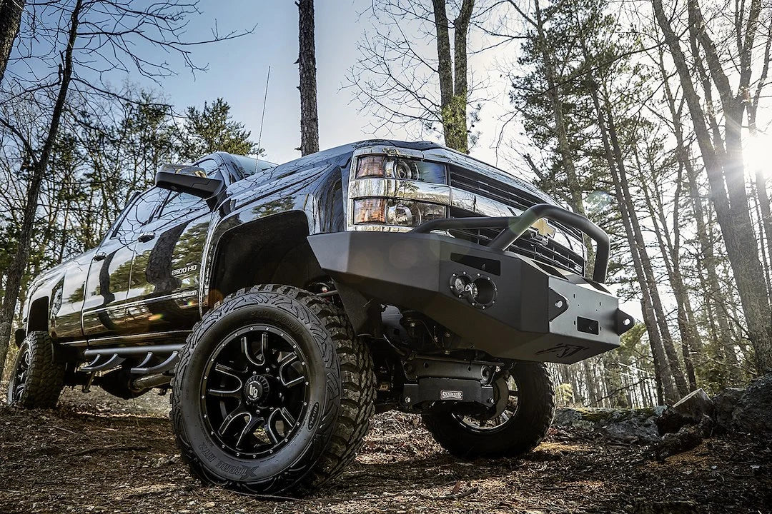 Low close-up of Fab Fours Premium heavy-duty winch front bumper with pre-runner guard on a lifted black Chevrolet Silverado 2500HD, passenger side, parked in the woods