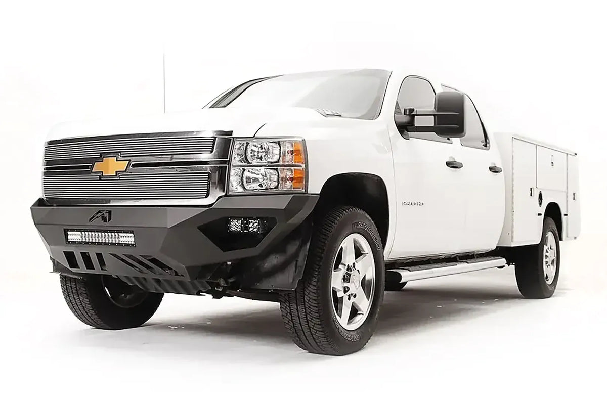 Fab Fours Vengeance front bumper on a white Chevrolet Silverado 2500HD, front driver-side three-quarter view, showing the angular bumper face, cube light pods, center light bar opening and skid plate