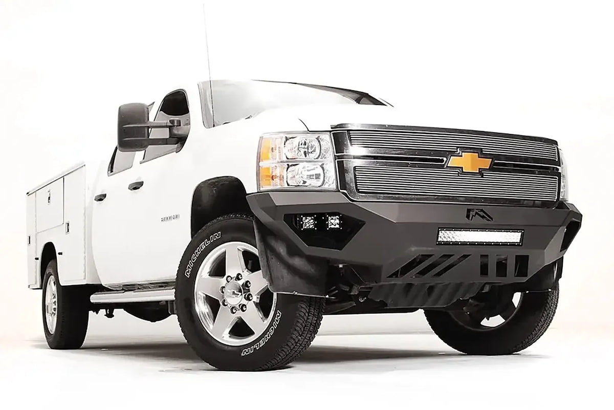Fab Fours Vengeance front bumper on a white Chevrolet Silverado 2500HD, front passenger-side three-quarter view, showing the angular bumper face, cube light pods, center light bar opening and skid plate