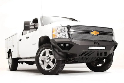 Fab Fours Vengeance front bumper on a white Chevrolet Silverado 2500HD, front passenger-side three-quarter view, showing the angular bumper face, cube light pods, center light bar opening and skid plate