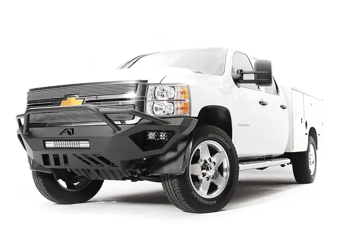 Fab Fours Vengeance front bumper with pre-runner guard on a white Chevrolet Silverado 2500HD, front driver-side three-quarter view, showing the pre-runner guard, cube light pods, center light bar opening and skid plate