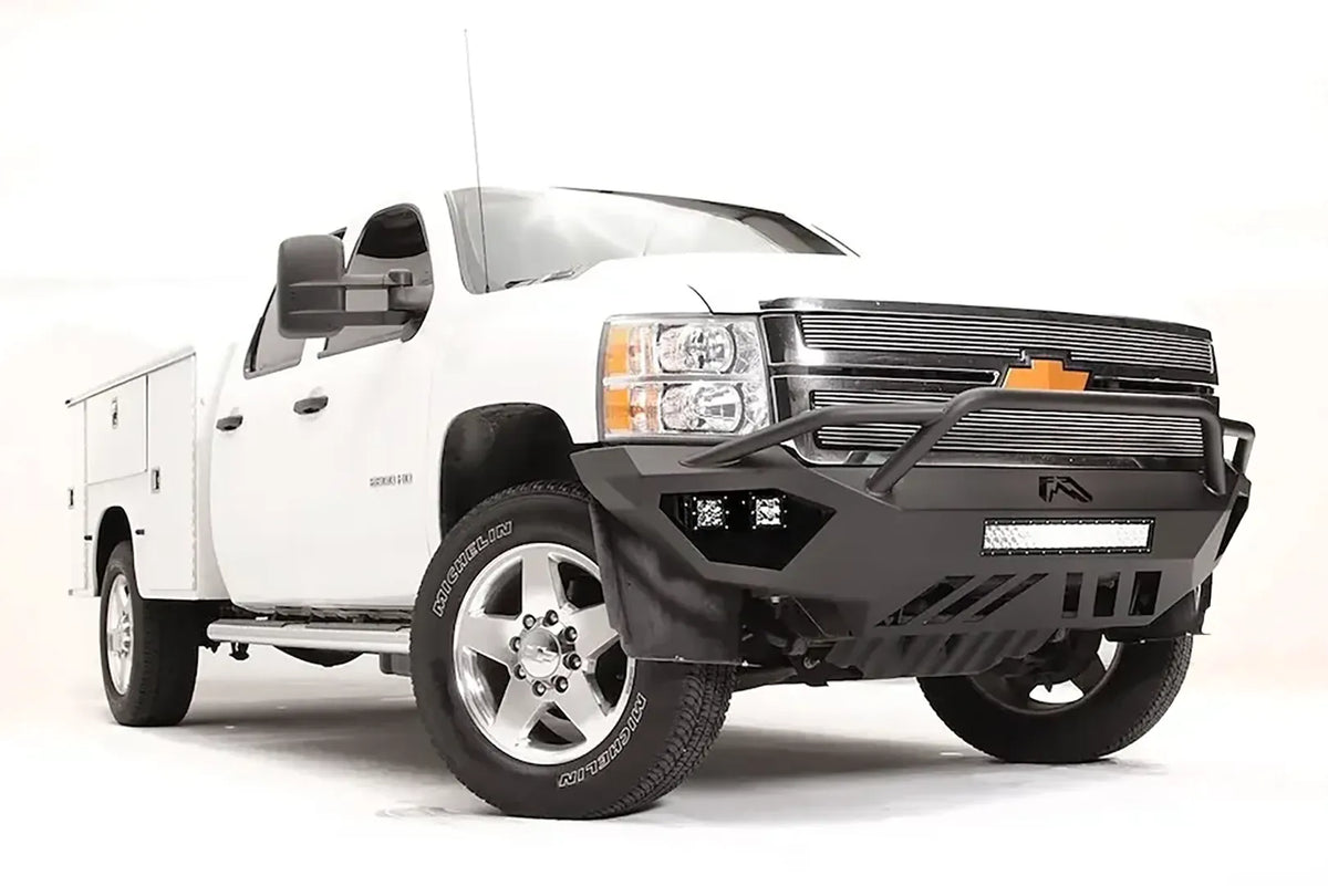Fab Fours Vengeance front bumper with pre-runner guard on a white Chevrolet Silverado 2500HD, front passenger-side three-quarter view, showing the pre-runner guard, cube light pods, center light bar opening and skid plate