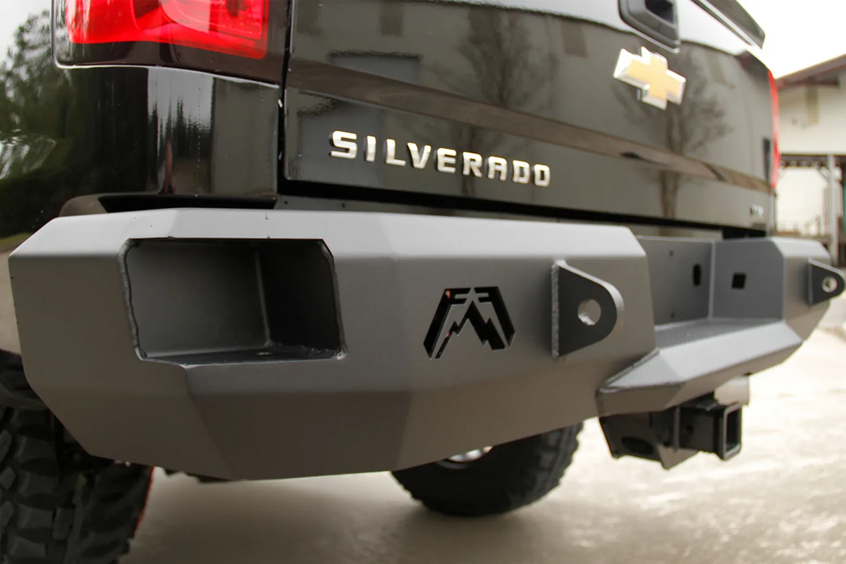 Fab Fours Premium rear bumper on a black Chevrolet Silverado 2500HD, close-up rear corner outdoor view, showing the Fab Fours logo cutout, shackle mount, square light opening and hitch receiver