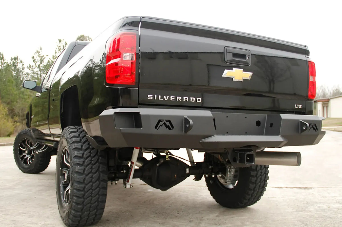 Fab Fours Premium rear bumper on a black Chevrolet Silverado 2500HD, rear driver-side three-quarter outdoor view, showing the Fab Fours logo cutouts, shackle mounts and angled bumper ends