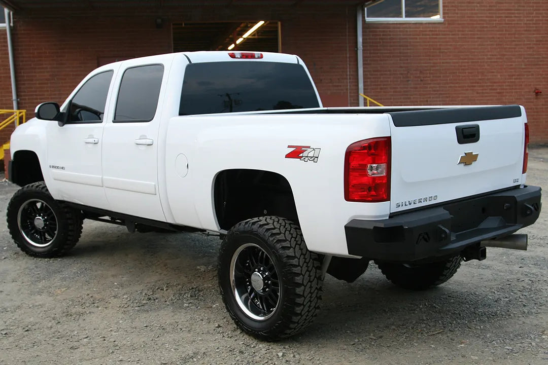 Fab Fours Premium rear bumper on a white Chevrolet Silverado 2500HD, rear driver-side three-quarter outdoor view, showing the angular bumper ends, Fab Fours logo cutouts and center step