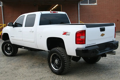 Fab Fours Premium rear bumper on a white Chevrolet Silverado 2500HD, rear driver-side three-quarter outdoor view, showing the angular bumper ends, Fab Fours logo cutouts and center step