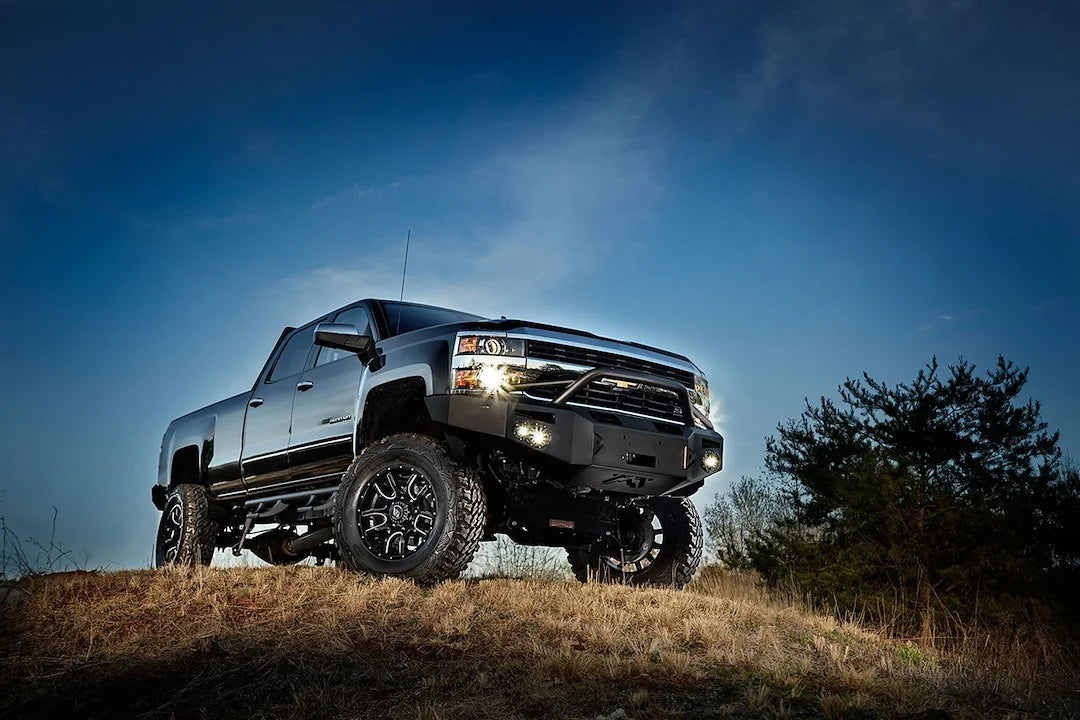 Lifted black Chevrolet Silverado 2500HD with Fab Fours Premium heavy-duty winch front bumper and pre-runner guard on a hilltop at dusk, fog lights on
