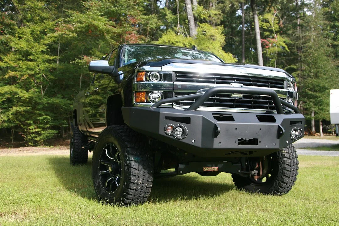 Fab Fours Premium Winch front bumper with pre-runner guard on a black Chevrolet Silverado 2500HD, front outdoor view, showing the pre-runner guard, center winch plate, round lights and shackle mounts