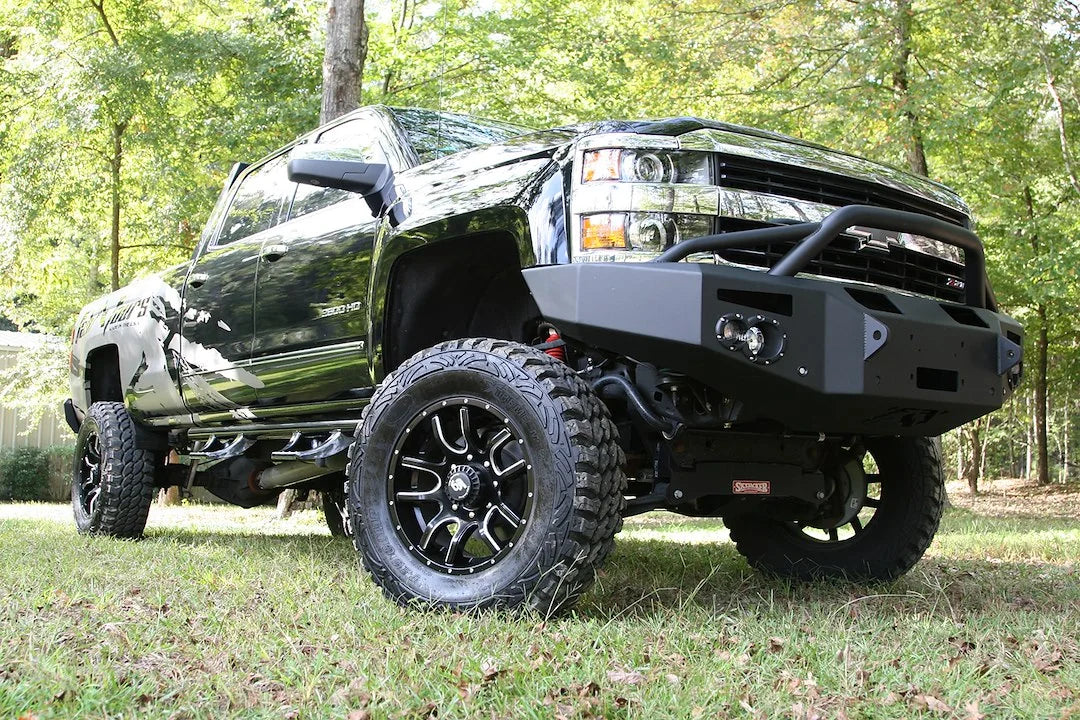 Fab Fours Premium Winch front bumper with pre-runner guard on a black Chevrolet Silverado 2500HD, front passenger-side three-quarter outdoor view, showing the pre-runner guard, center winch plate and round lights