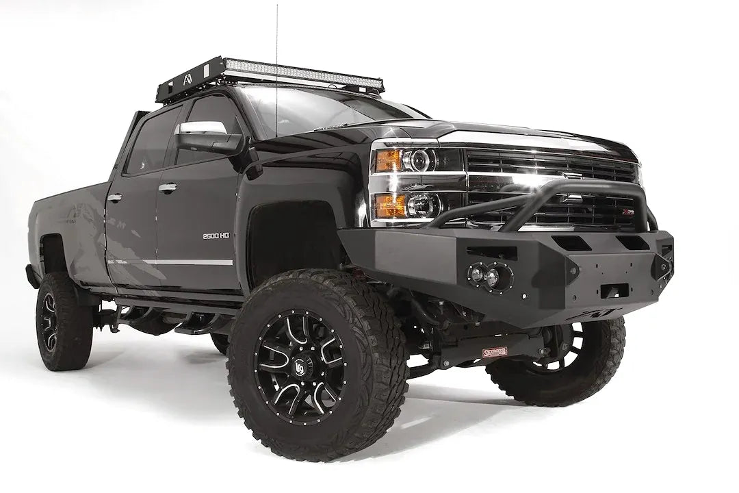 Fab Fours Premium Winch front bumper with pre-runner guard on a black Chevrolet Silverado 2500HD, front passenger-side three-quarter view, showing the pre-runner guard, center winch plate, round lights and shackle mounts