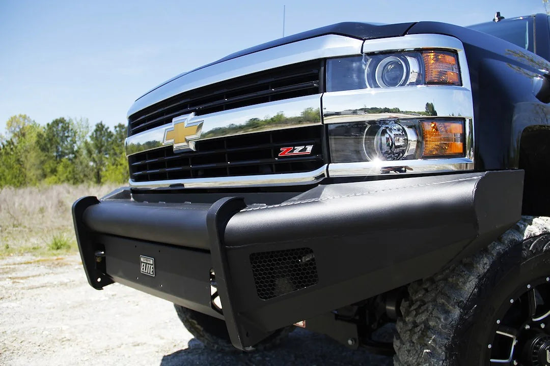 Fab Fours Black Steel Elite front bumper on a black Chevrolet Silverado 2500HD, close-up front corner view, showing the smooth-plate bumper face, Elite badge and mesh light opening