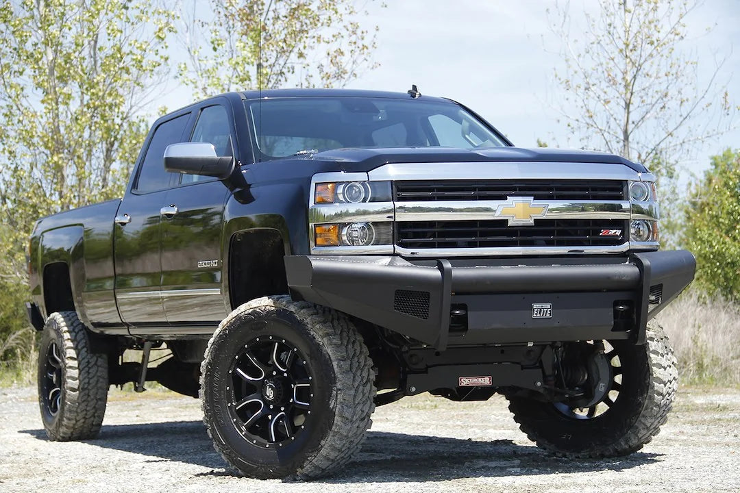 Fab Fours Black Steel Elite front bumper on a black Chevrolet Silverado 2500HD, front passenger-side three-quarter outdoor view, showing the smooth-plate bumper face, Elite badge and mesh light openings
