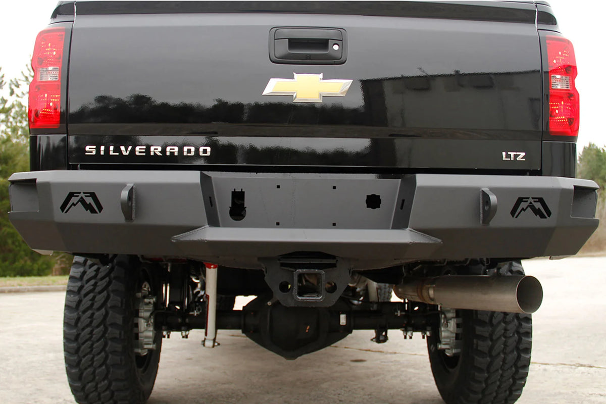 Fab Fours Premium rear bumper on a black Chevrolet Silverado, straight-on rear view outdoors, showing the cut-out Fab Fours logos, the D-ring shackle mounts, the center license plate panel and the hitch receiver opening