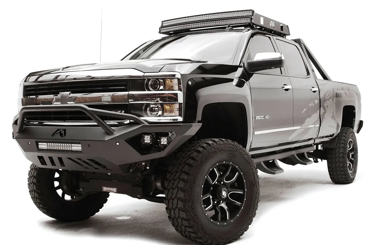 Fab Fours Vengeance front bumper with pre-runner guard on a black Chevrolet Silverado 2500HD, front driver-side three-quarter view, showing the pre-runner guard, cube light pods, center light bar and skid plate