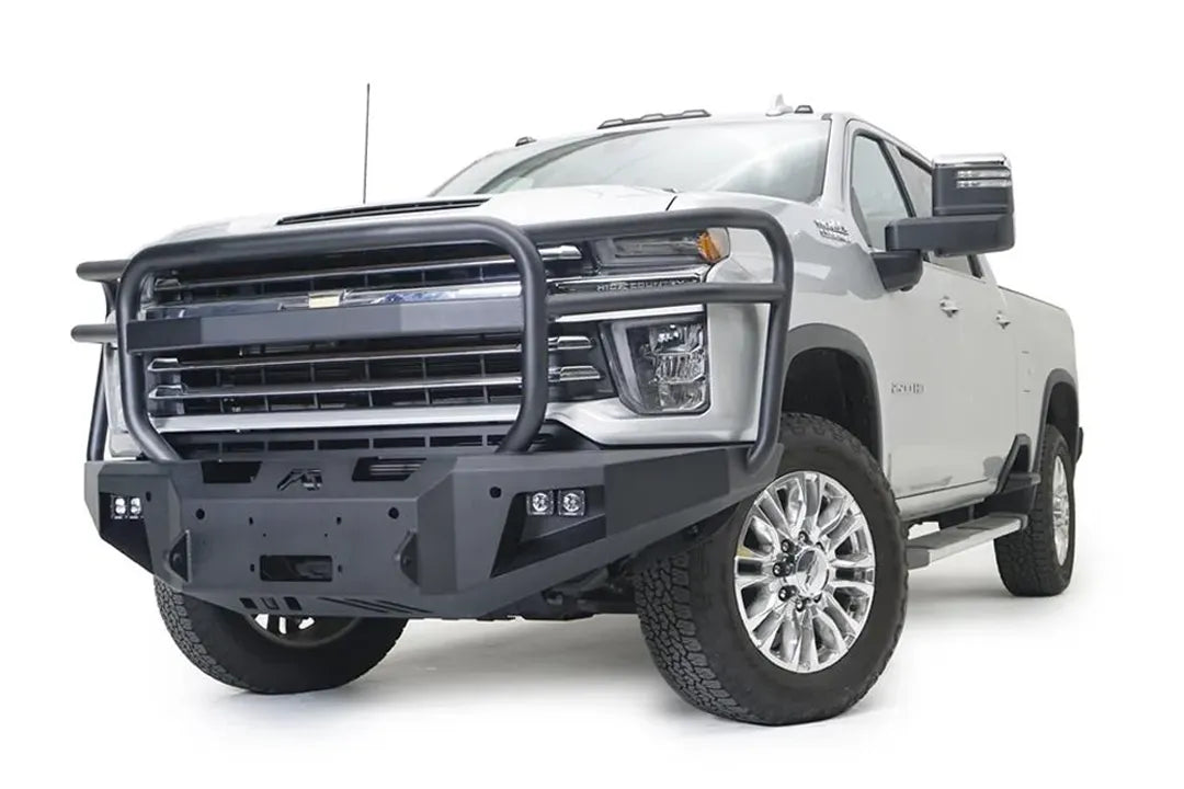 Fab Fours Premium Winch front bumper with full guard on a white Chevrolet Silverado 2500HD, front driver-side three-quarter view, showing the full grille guard, the center winch plate, cube light pods and Fab Fours logo