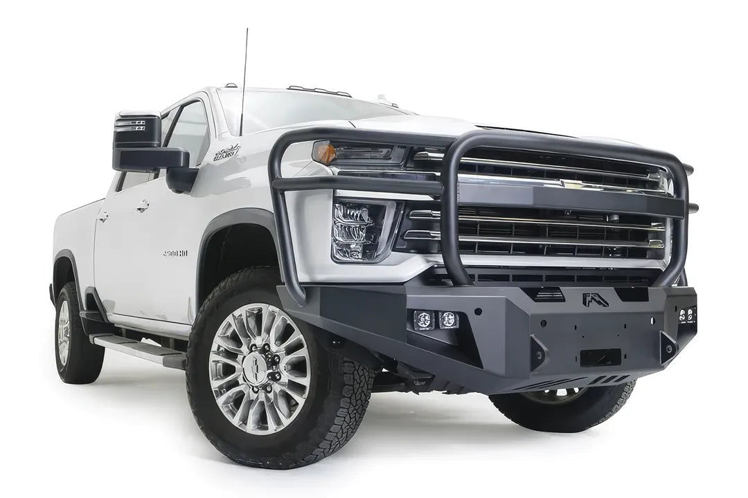Fab Fours Premium Winch front bumper with full guard on a white Chevrolet Silverado 2500HD, front passenger-side three-quarter view, showing the full grille guard, the center winch plate, cube light pods and angular bumper ends