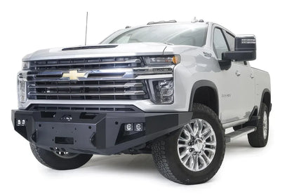 Fab Fours Premium Winch front bumper on a white Chevrolet Silverado 2500HD, front driver-side three-quarter view, showing the center winch plate, cube light pods and Fab Fours logo