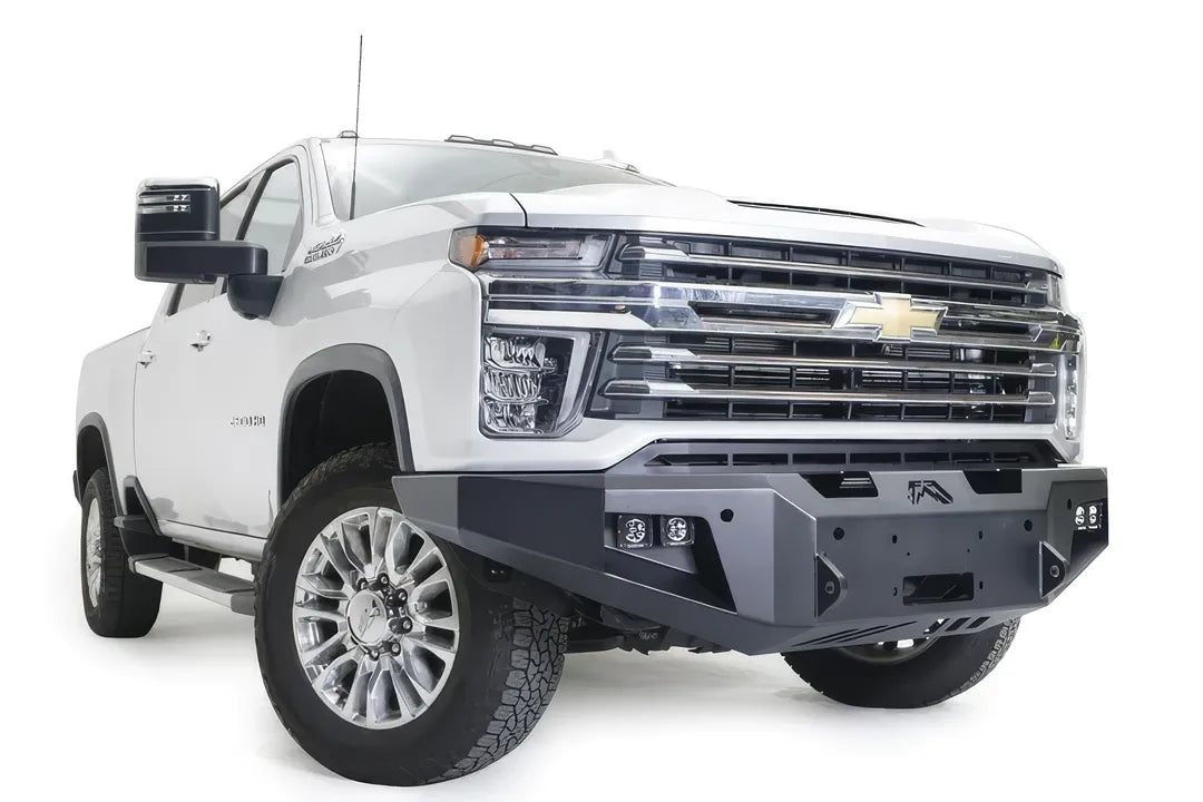 Fab Fours Premium Winch front bumper on a white Chevrolet Silverado 2500HD, front passenger-side three-quarter view, showing the center winch plate, cube light pods and angular bumper ends