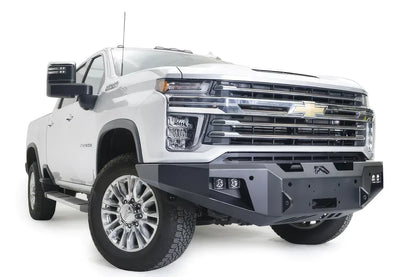 Fab Fours Premium Winch front bumper on a white Chevrolet Silverado 2500HD, front passenger-side three-quarter view, showing the center winch plate, cube light pods and angular bumper ends