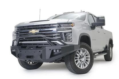 Fab Fours Premium Winch front bumper with pre-runner guard on a white Chevrolet Silverado 2500HD, front driver-side three-quarter view, showing the pre-runner guard, the center winch plate, cube light pods and Fab Fours logo
