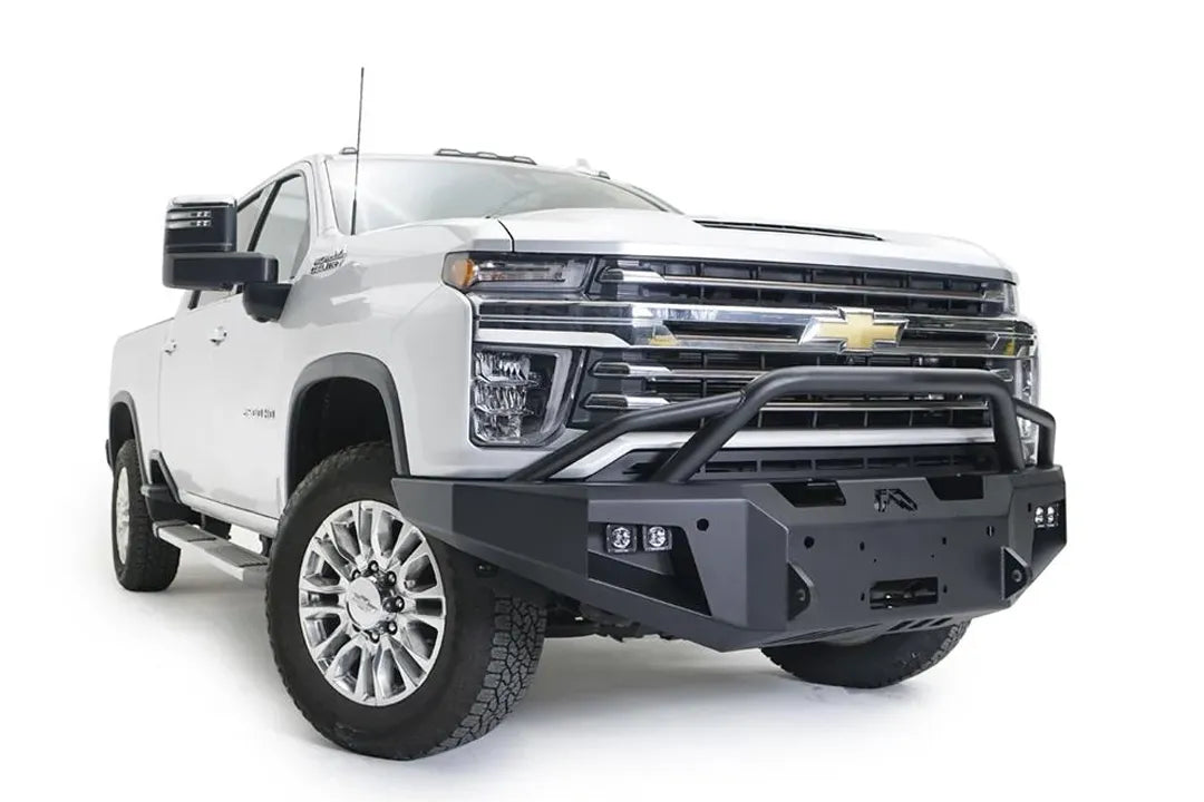Fab Fours Premium Winch front bumper with pre-runner guard on a white Chevrolet Silverado 2500HD, front passenger-side three-quarter view, showing the pre-runner guard, the center winch plate, cube light pods and angular bumper ends