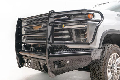 Fab Fours Black Steel front bumper with full guard on a white Chevrolet Silverado 2500HD, close-up of the front corner, showing the mesh-filled guard, diamond-plate end cap and lower skid slots