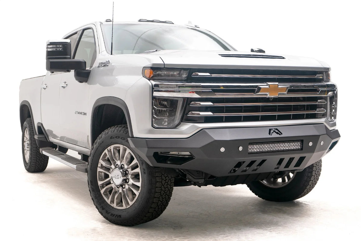 Fab Fours Vengeance front bumper on a white Chevrolet Silverado 2500HD, low front passenger-side three-quarter view, showing the angular bumper wings, center light bar and skid plate cutouts