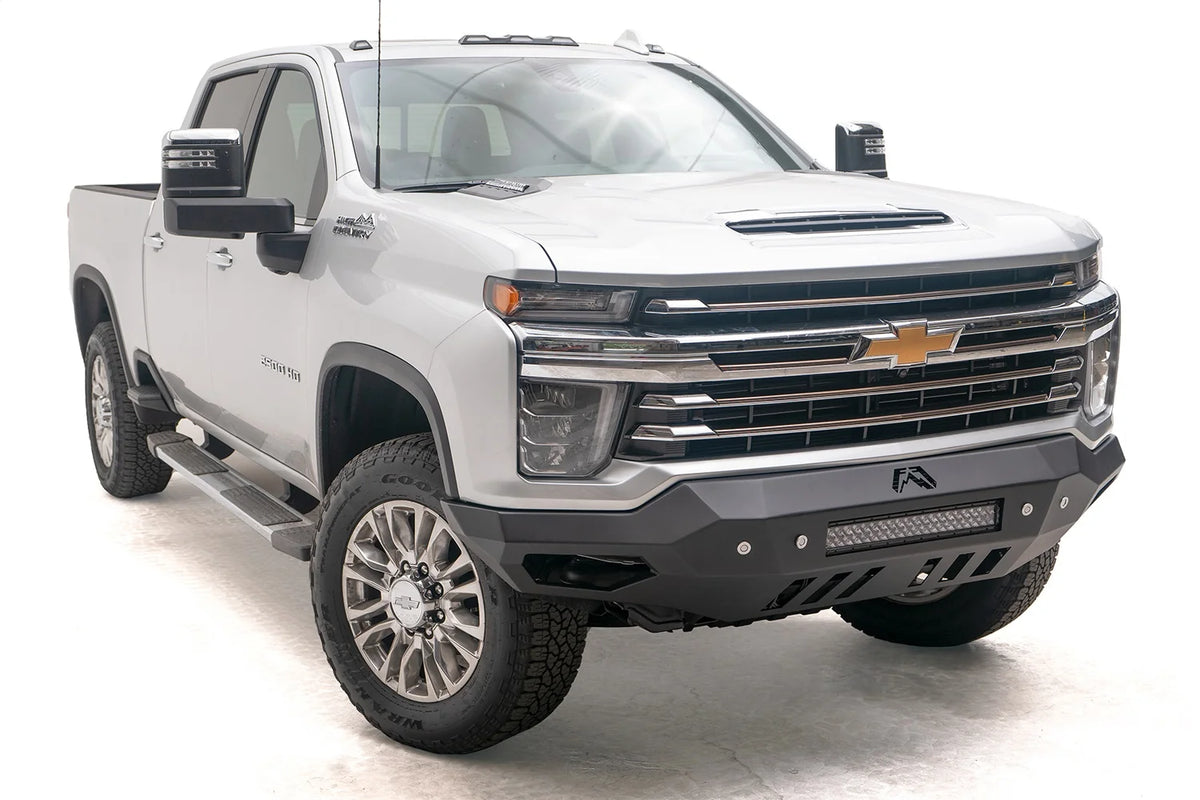 Fab Fours Vengeance front bumper on a white Chevrolet Silverado 2500HD, front passenger-side three-quarter view, showing the no-guard design, integrated light bar and slotted lower skid plate
