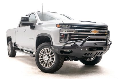 Fab Fours Vengeance front bumper with pre-runner guard on a white Chevrolet Silverado 2500HD, low front passenger-side three-quarter view, showing the tubular pre-runner hoop, angular end wings and slotted skid plate