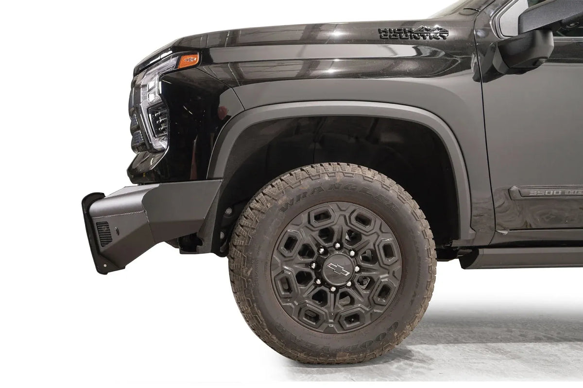 Fab Fours Black Steel Elite front bumper on a black Chevrolet Silverado 2500HD, side close-up of the front corner, showing the bumper end wing, mesh vent and wheel clearance