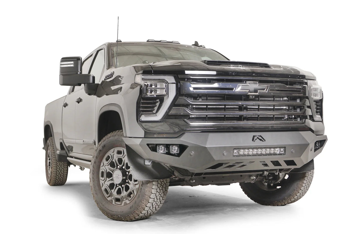 Fab Fours Vengeance front bumper on a dark gray Chevrolet Silverado 2500HD, front passenger-side three-quarter view, showing the integrated light bar, pod light openings and slotted skid plate