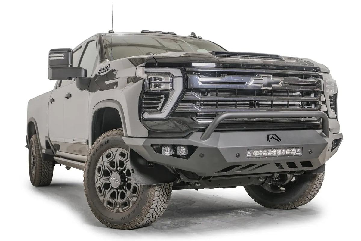 Fab Fours Vengeance front bumper with pre-runner guard on a dark gray Chevrolet Silverado 2500HD, front passenger-side three-quarter view, showing the tubular pre-runner hoop, integrated light bar and pod light openings