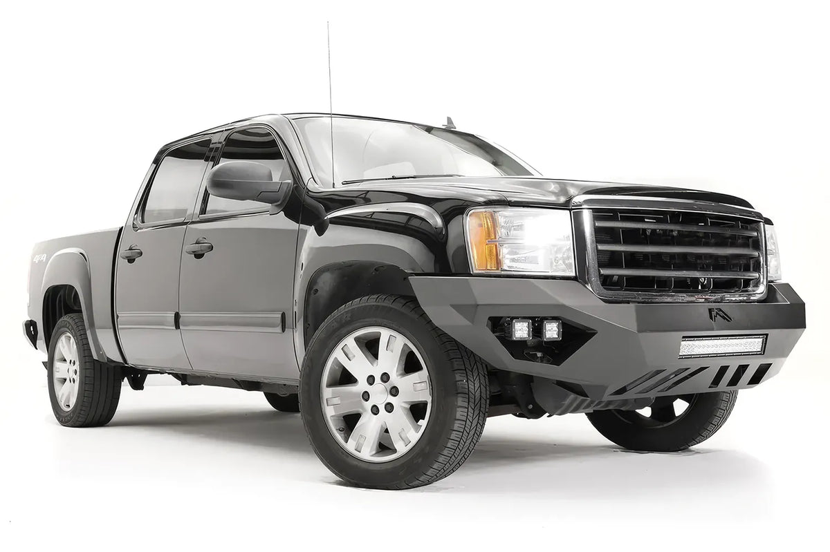 Fab Fours Vengeance front bumper on a black GMC Sierra 1500, front passenger-side three-quarter view, showing the angular end wings, cube light pods and slotted skid plate
