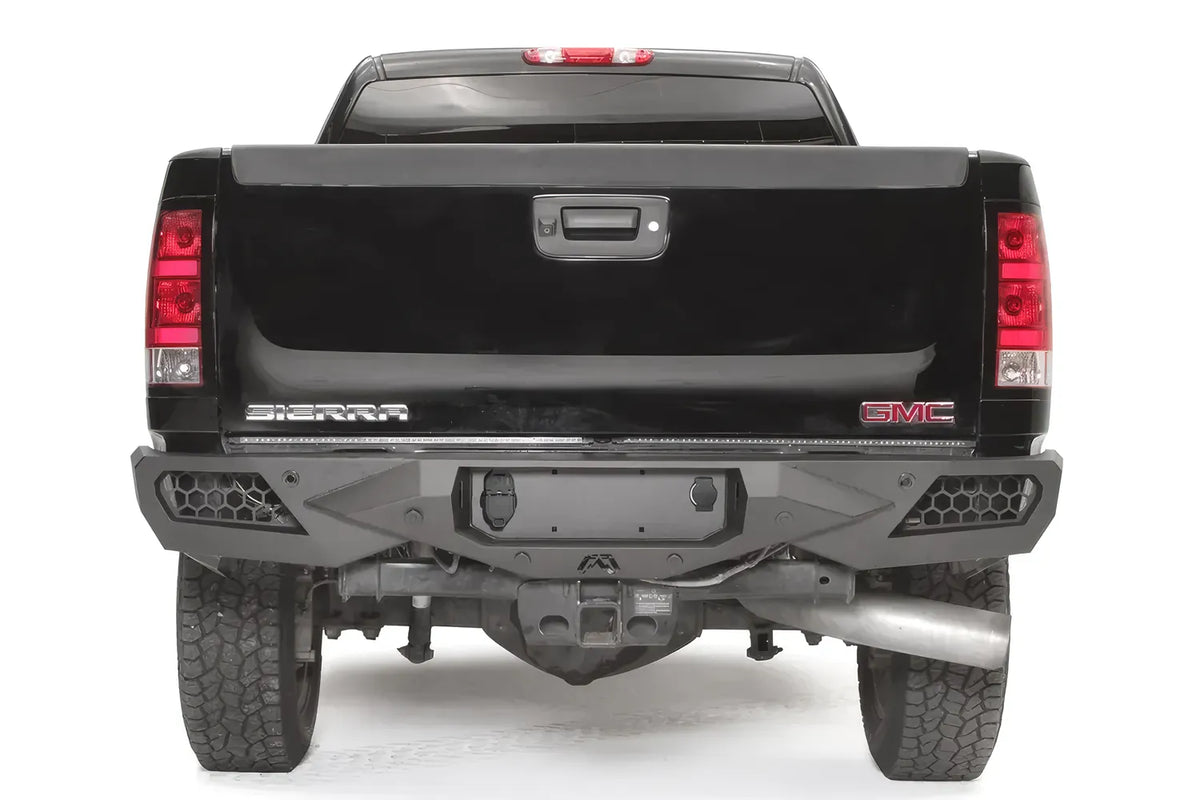 Fab Fours Vengeance rear bumper on a black GMC Sierra 1500, straight-on rear view, showing the angular black steel face, honeycomb mesh end vents and center hitch area