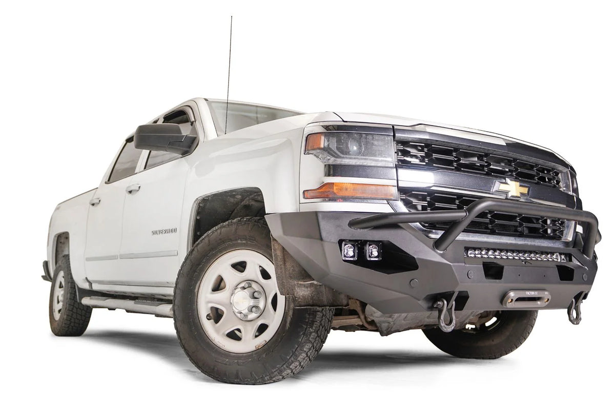 Fab Fours Matrix Front Bumper with pre-runner guard on a white Chevrolet Silverado 1500, front passenger-side view, showing the tubular guard over the LED light bar, dual cube lights, winch fairlead and D-ring shackles
