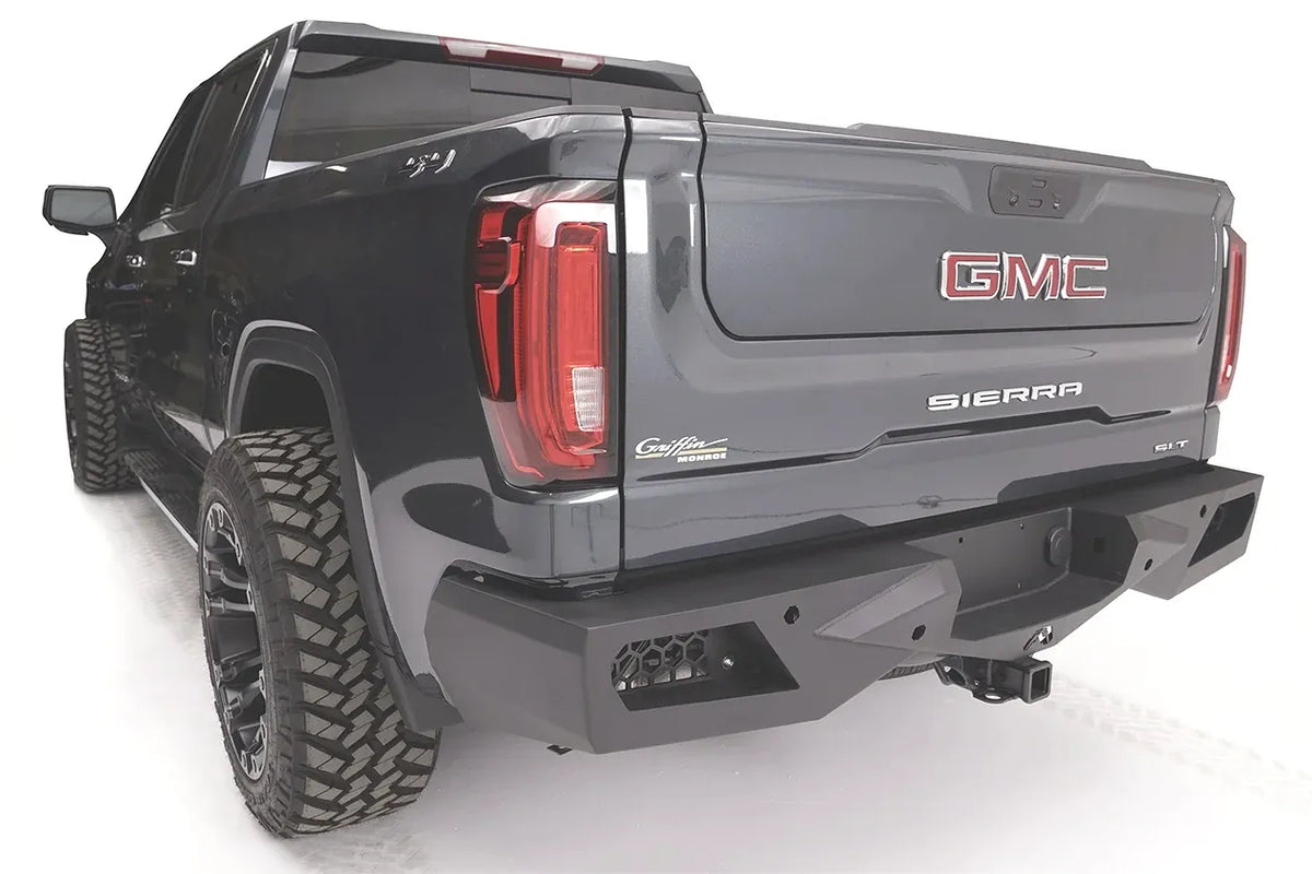 Fab Fours Vengeance rear bumper on a gray GMC Sierra 1500, rear driver-side three-quarter view, showing the angular end cap, honeycomb light pockets, and hitch receiver