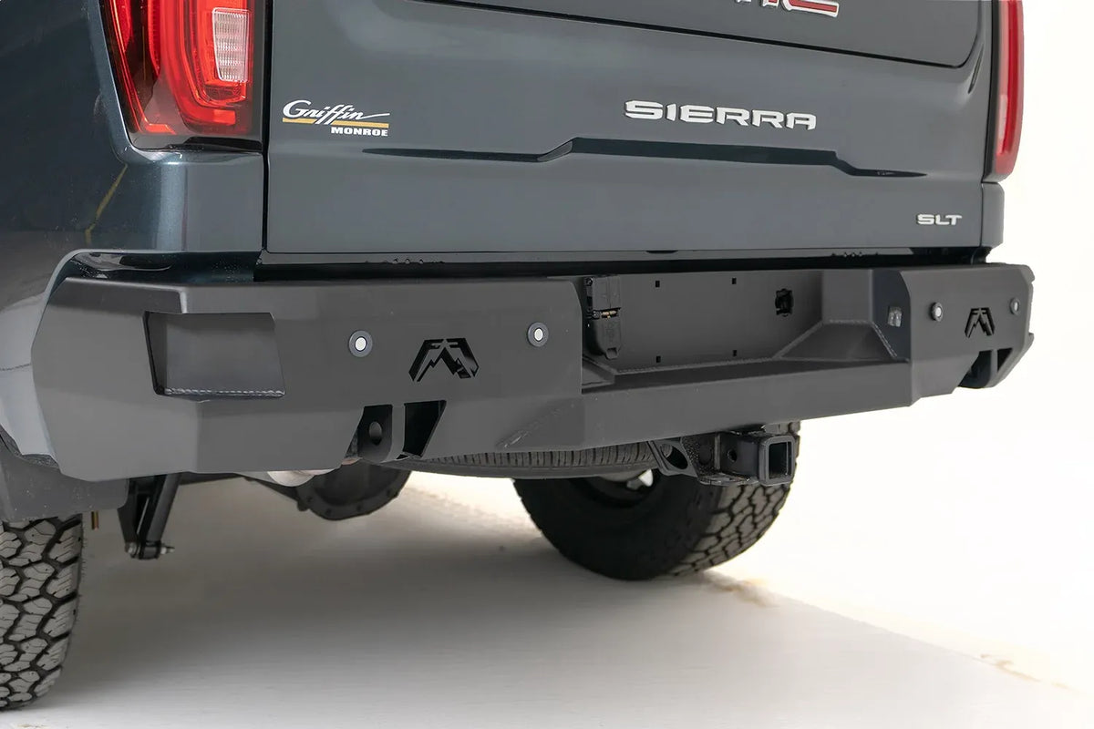 Fab Fours Premium rear bumper on a gray GMC Sierra 1500, close rear three-quarter view, showing the plate recess, end-cap logos, and hitch receiver
