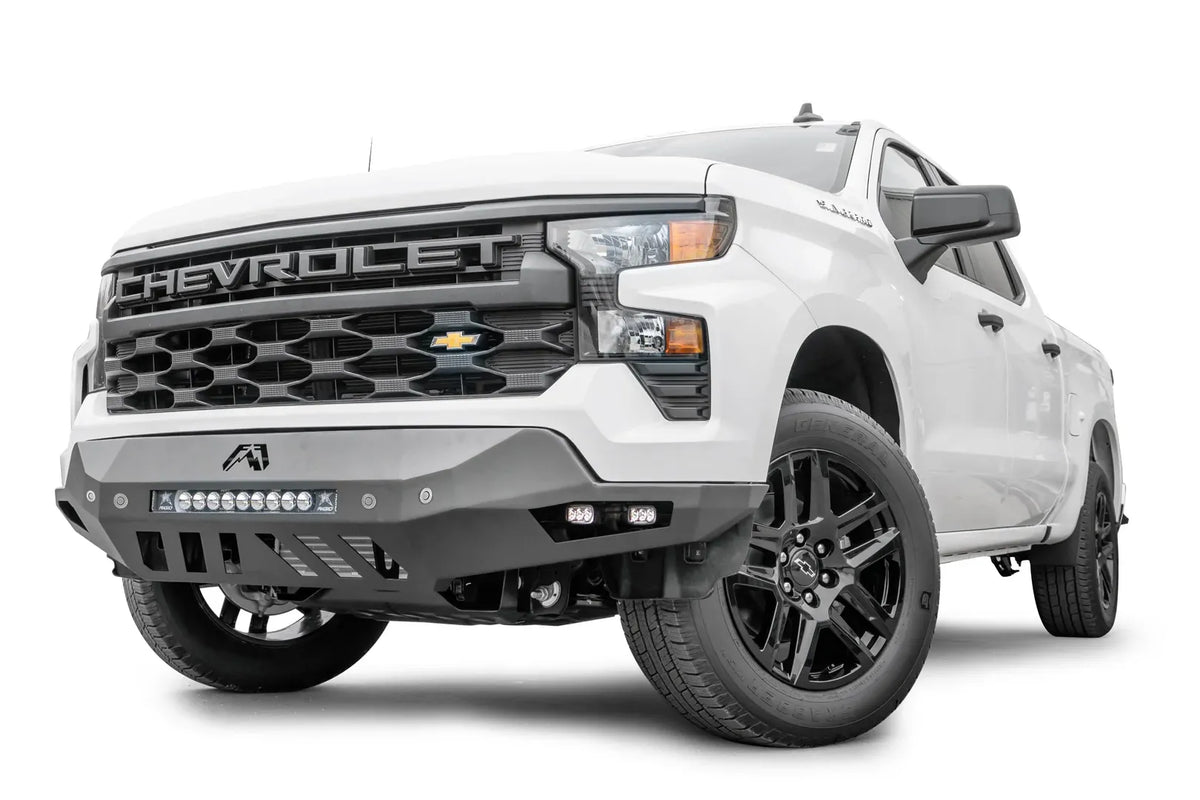 Fab Fours Vengeance front bumper on a white Chevrolet Silverado 1500, front driver-side three-quarter view, showing the LED light bar, cube light pods, and skid plate