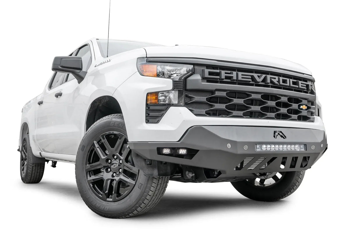 Fab Fours Vengeance front bumper on a white Chevrolet Silverado 1500, front passenger-side three-quarter view, showing the angular bumper wing, LED light bar, and slotted skid plate