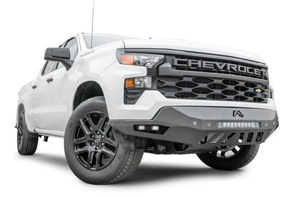 Fab Fours Vengeance front bumper on a white Chevrolet Silverado 1500, front passenger-side three-quarter view, showing the angular bumper wing, LED light bar, and slotted skid plate