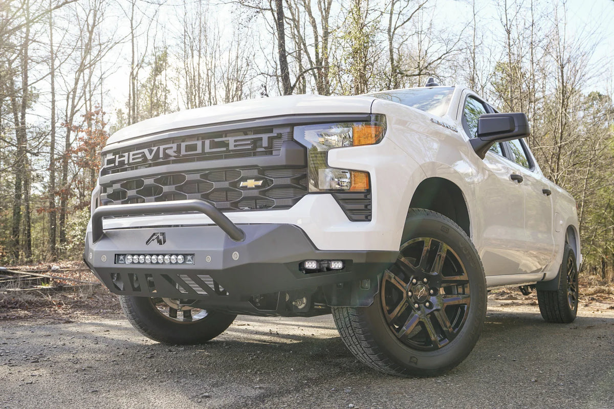 Fab Fours Vengeance front bumper with pre-runner guard on a white Chevrolet Silverado 1500, front driver-side three-quarter view outdoors, showing the pre-runner bar, LED light bar, and slotted skid plate