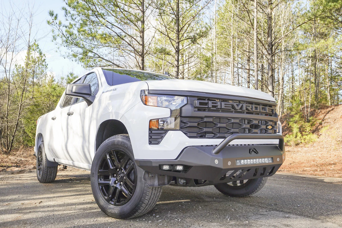 Fab Fours Vengeance front bumper with pre-runner guard on a white Chevrolet Silverado 1500, front passenger-side three-quarter view outdoors, showing the pre-runner bar, angular bumper wing, and skid plate